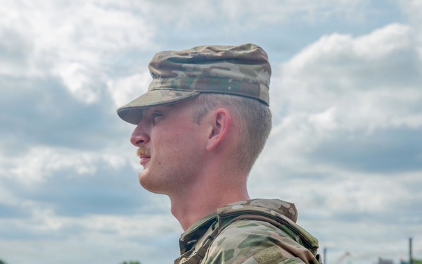 A male soldier in U.S. Army Operational Camouflage Pattern (OCP) uniform stands in profile view near a waterway, appearing to observe the surroundings. He wears a patrol cap and a shoulder patch depicting a shield with a sun/star design, consistent with U.S. Army unit insignia. The background shows a calm river or harbor with docks, trees, and overcast skies.