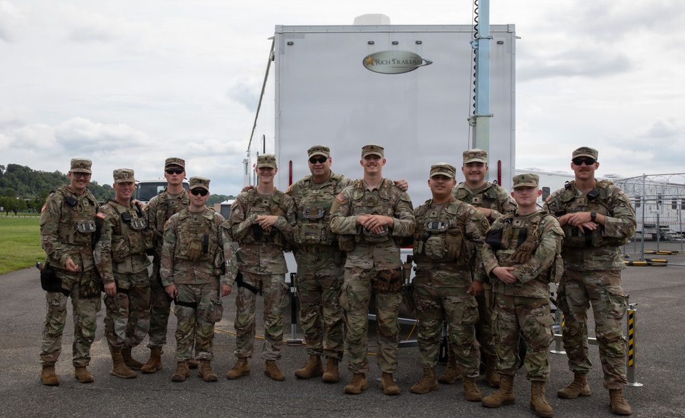 A group of approximately eleven military personnel in matching camouflage uniforms pose together in front of a white 'Rich Trailers' trailer. They appear to be in a relaxed, informal group photo taken outdoors on a paved surface. The setting suggests a military event, exercise, or security detail deployment.