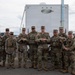 A group of approximately eleven military personnel in matching camouflage uniforms pose together in front of a white 'Rich Trailers' trailer. They appear to be in a relaxed, informal group photo taken outdoors on a paved surface. The setting suggests a military event, exercise, or security detail deployment.