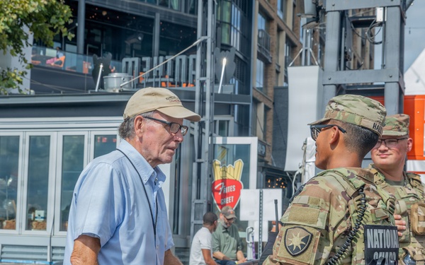 An older civilian man wearing a tan cap, glasses, and a light blue shirt is conversing with two U.S. National Guard soldiers in camouflage uniforms at what appears to be an outdoor public event or festival. The soldiers are wearing tactical vests with visible 'National Guard' patches and unit insignia. The setting includes metal crowd-control barriers, vendor structures, and urban buildings in the background.