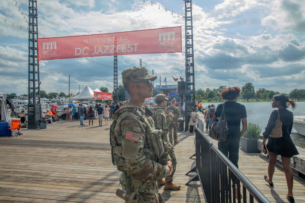 The image shows military personnel in U.S. Army camouflage uniforms providing security at the DC JazzFest, a public music festival held on a waterfront boardwalk. A prominent red banner reading 'Welcome to DC JazzFest' is visible overhead, with festival-goers and event infrastructure in the background. The setting appears to be a marina or waterfront venue with boats visible on the left side.