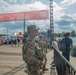 The image shows military personnel in U.S. Army camouflage uniforms providing security at the DC JazzFest, a public music festival held on a waterfront boardwalk. A prominent red banner reading 'Welcome to DC JazzFest' is visible overhead, with festival-goers and event infrastructure in the background. The setting appears to be a marina or waterfront venue with boats visible on the left side.