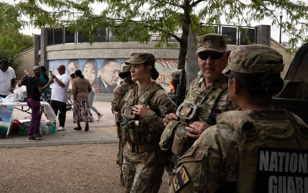 National Guard personnel in camouflage uniforms are present at an outdoor public gathering or community event. At least three soldiers are visible in the foreground, one bearing an 'MP' (Military Police) patch and another identified by a 'JACINTO' name tape. In the background, civilians are gathered near tables in front of a mural depicting what appears to be Dr. Martin Luther King Jr.