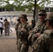National Guard personnel in camouflage uniforms are present at an outdoor public gathering or community event. At least three soldiers are visible in the foreground, one bearing an 'MP' (Military Police) patch and another identified by a 'JACINTO' name tape. In the background, civilians are gathered near tables in front of a mural depicting what appears to be Dr. Martin Luther King Jr.