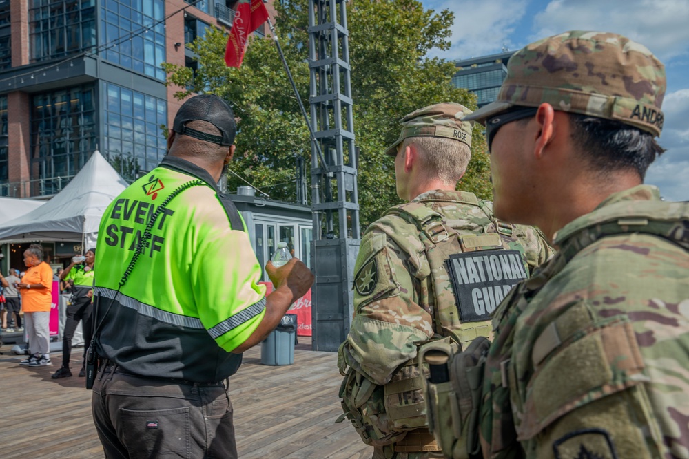 Three individuals are standing on a wooden deck or boardwalk at an outdoor event in an urban setting. Two are National Guard soldiers in full camouflage uniforms and tactical gear, and one is an event staff member wearing a high-visibility yellow vest. They appear to be coordinating or communicating at what looks like a public event.