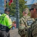 Three individuals are standing on a wooden deck or boardwalk at an outdoor event in an urban setting. Two are National Guard soldiers in full camouflage uniforms and tactical gear, and one is an event staff member wearing a high-visibility yellow vest. They appear to be coordinating or communicating at what looks like a public event.