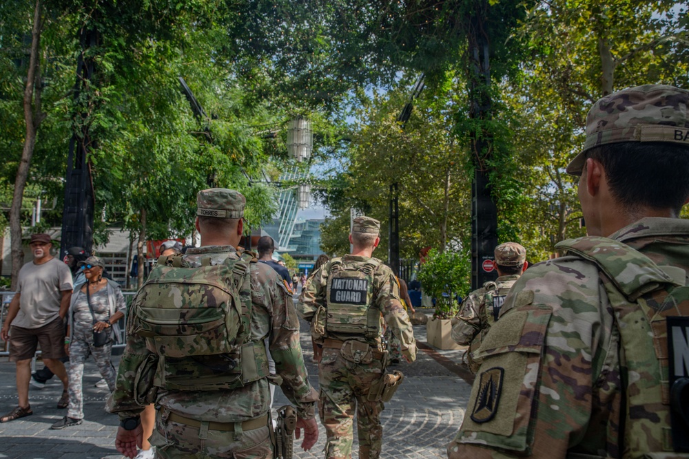 Several U.S. National Guard soldiers in full camouflage uniforms and tactical gear are walking through a public outdoor area, likely a park or plaza in an urban setting. The soldiers are moving away from the camera, with 'NATIONAL GUARD' clearly visible on the back of one soldier's vest. Civilians are also present in the background, suggesting a public security or patrol deployment.