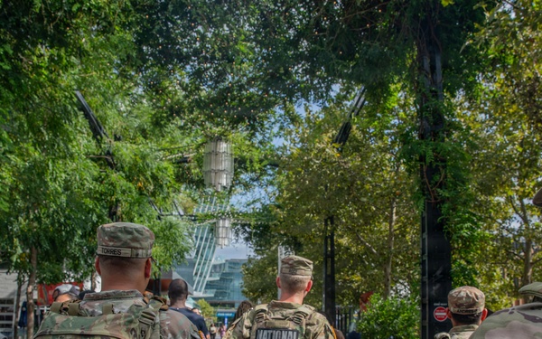 Several U.S. National Guard soldiers in full camouflage uniforms and tactical gear are walking through a public outdoor area, likely a park or plaza in an urban setting. The soldiers are moving away from the camera, with 'NATIONAL GUARD' clearly visible on the back of one soldier's vest. Civilians are also present in the background, suggesting a public security or patrol deployment.
