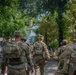 Several U.S. National Guard soldiers in full camouflage uniforms and tactical gear are walking through a public outdoor area, likely a park or plaza in an urban setting. The soldiers are moving away from the camera, with 'NATIONAL GUARD' clearly visible on the back of one soldier's vest. Civilians are also present in the background, suggesting a public security or patrol deployment.