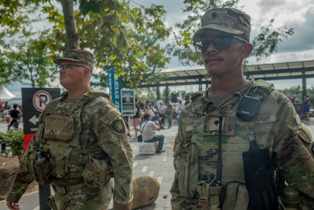 Two U.S. Army soldiers in OCP (Operational Camouflage Pattern) uniforms are standing at what appears to be a public outdoor event or venue. The foreground soldier is wearing a tactical vest with a radio and has 'ARMY' tape visible on his uniform. They appear to be providing security or a visible military presence at a civilian gathering.