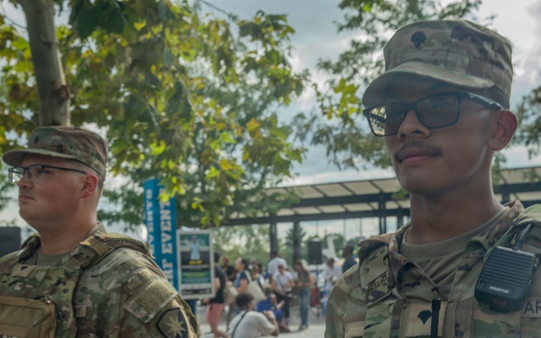 Two U.S. Army soldiers in OCP (Operational Camouflage Pattern) uniforms are standing at what appears to be a public outdoor event or venue. The foreground soldier is wearing a tactical vest with a radio and has 'ARMY' tape visible on his uniform. They appear to be providing security or a visible military presence at a civilian gathering.
