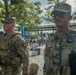 Two U.S. Army soldiers in OCP (Operational Camouflage Pattern) uniforms are standing at what appears to be a public outdoor event or venue. The foreground soldier is wearing a tactical vest with a radio and has 'ARMY' tape visible on his uniform. They appear to be providing security or a visible military presence at a civilian gathering.