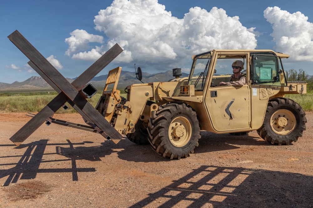 A military service member operates a tan/desert-colored military forklift or rough-terrain telehandler on a dirt surface in what appears to be a southwestern U.S. landscape. The vehicle is carrying large X-shaped steel anti-vehicle barriers (commonly known as Czech hedgehogs or dragon's teeth) on its forks. The setting features arid terrain, distant mountains, and large cumulus clouds in a blue sky.