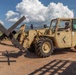 A military service member operates a tan/desert-colored military forklift or rough-terrain telehandler on a dirt surface in what appears to be a southwestern U.S. landscape. The vehicle is carrying large X-shaped steel anti-vehicle barriers (commonly known as Czech hedgehogs or dragon's teeth) on its forks. The setting features arid terrain, distant mountains, and large cumulus clouds in a blue sky.
