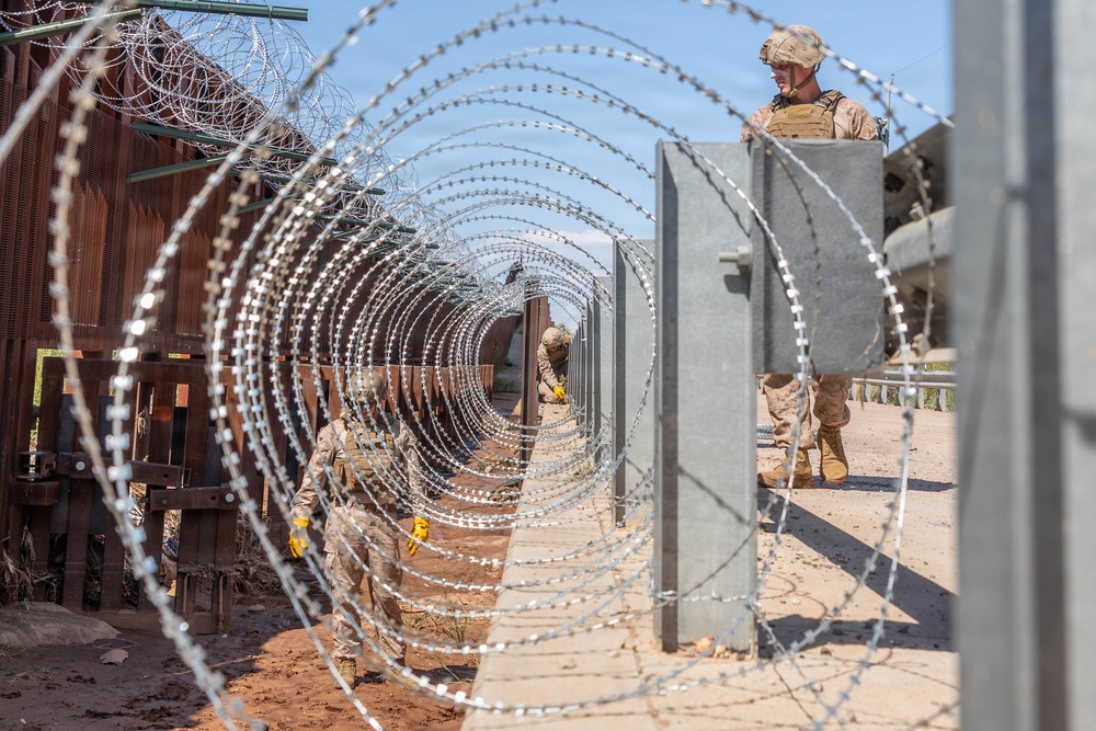Military personnel in camouflage uniforms and tactical gear are installing concertina (razor) wire along a border barrier consisting of rust-colored steel bollards. The soldiers are working between the existing fence and newly placed concrete jersey barriers, extending the wire in a coiled tunnel formation. The scene depicts an active border security fortification operation in an arid, desert environment.