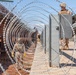 Military personnel in camouflage uniforms and tactical gear are installing concertina (razor) wire along a border barrier consisting of rust-colored steel bollards. The soldiers are working between the existing fence and newly placed concrete jersey barriers, extending the wire in a coiled tunnel formation. The scene depicts an active border security fortification operation in an arid, desert environment.