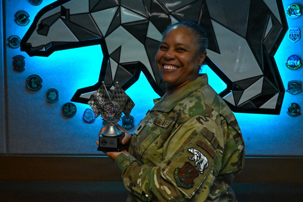 A U.S. Air Force service member in OCP (Operational Camouflage Pattern) uniform is smiling and holding a trophy with checkered flags and an Alaska-themed medallion. She is standing in front of a geometric bear wall art installation with blue LED backlighting, surrounded by military unit patches/coins mounted on the wall. The setting appears to be an official military facility, likely at a base in Alaska.