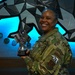 A U.S. Air Force service member in OCP (Operational Camouflage Pattern) uniform is smiling and holding a trophy with checkered flags and an Alaska-themed medallion. She is standing in front of a geometric bear wall art installation with blue LED backlighting, surrounded by military unit patches/coins mounted on the wall. The setting appears to be an official military facility, likely at a base in Alaska.