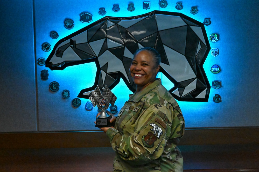 A U.S. Air Force service member in OCP (Operational Camouflage Pattern) uniform is smiling and holding a trophy or award. She is standing in front of a geometric polar bear wall art installation with blue LED backlighting, surrounded by military unit patches/coins. The patch on her shoulder appears to be associated with Alaskan Command or a unit based in Alaska.