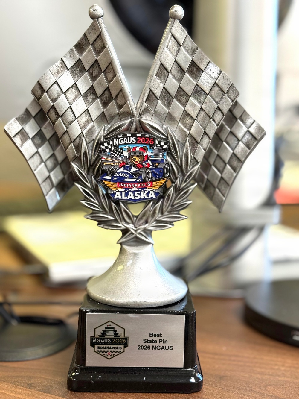 A silver racing-themed trophy featuring two crossed checkered flags and a colorful center medallion. The trophy was awarded for 'Best State Pin' at the 2026 NGAUS (National Guard Association of the United States) conference in Indianapolis. The Alaska state delegation won the award, as indicated by the Alaska-themed pin displayed on the trophy's center medallion.