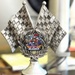 A silver racing-themed trophy featuring two crossed checkered flags and a colorful center medallion. The trophy was awarded for 'Best State Pin' at the 2026 NGAUS (National Guard Association of the United States) conference in Indianapolis. The Alaska state delegation won the award, as indicated by the Alaska-themed pin displayed on the trophy's center medallion.