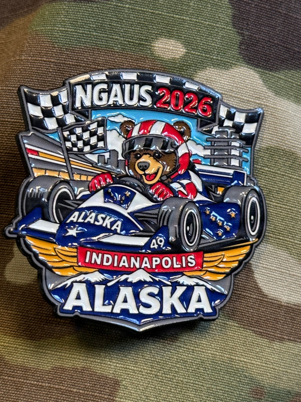 A colorful enamel challenge coin or lapel pin representing Alaska's delegation to the NGAUS 2026 conference in Indianapolis. The pin features a bear mascot wearing a racing helmet driving an IndyCar decorated with Alaska state flag imagery, combining Indianapolis Motor Speedway racing themes with Alaskan symbolism. The pin is photographed on a camouflage fabric background.