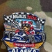 A colorful enamel challenge coin or lapel pin representing Alaska's delegation to the NGAUS 2026 conference in Indianapolis. The pin features a bear mascot wearing a racing helmet driving an IndyCar decorated with Alaska state flag imagery, combining Indianapolis Motor Speedway racing themes with Alaskan symbolism. The pin is photographed on a camouflage fabric background.
