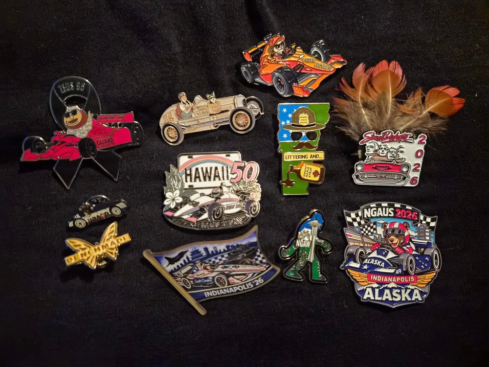 The image shows a collection of approximately 13 decorative enamel lapel pins arranged on a dark black fabric background. The pins are predominantly racing and Indianapolis 500-themed, featuring various states, characters, and NGAUS (National Guard Association of the United States) branding. A few loose feathers are also visible in the upper right corner of the image.