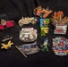 The image shows a collection of approximately 13 decorative enamel lapel pins arranged on a dark black fabric background. The pins are predominantly racing and Indianapolis 500-themed, featuring various states, characters, and NGAUS (National Guard Association of the United States) branding. A few loose feathers are also visible in the upper right corner of the image.