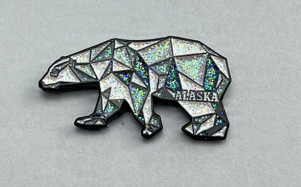 The image shows a decorative souvenir magnet or pin in the shape of a polar bear with a geometric, low-poly design. The bear's body is filled with holographic glitter segments separated by black outlines, creating a mosaic-like appearance. The word 'ALASKA' is embossed in silver lettering on the bear's body.