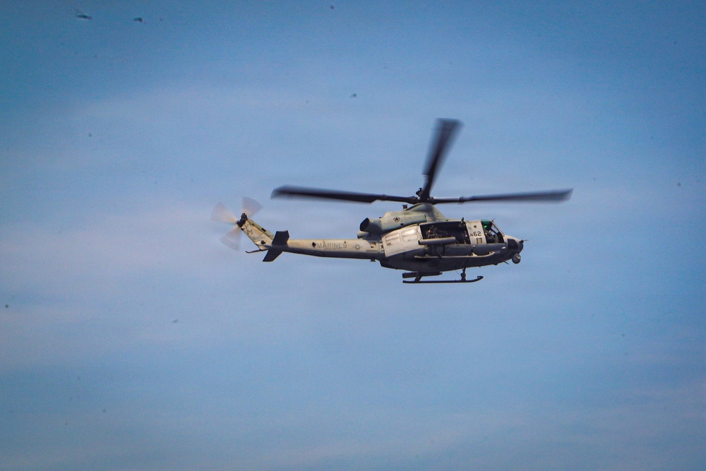 A U.S. Marines military helicopter is captured in flight against a blue sky with light cloud cover. The aircraft appears to be a Bell UH-1Y Venom or similar variant, identifiable by the 'MARINES' marking on the fuselage and the number '62' near the cockpit. The rotors are in motion, visible as a blur, indicating active flight.