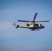 A U.S. Marines military helicopter is captured in flight against a blue sky with light cloud cover. The aircraft appears to be a Bell UH-1Y Venom or similar variant, identifiable by the 'MARINES' marking on the fuselage and the number '62' near the cockpit. The rotors are in motion, visible as a blur, indicating active flight.