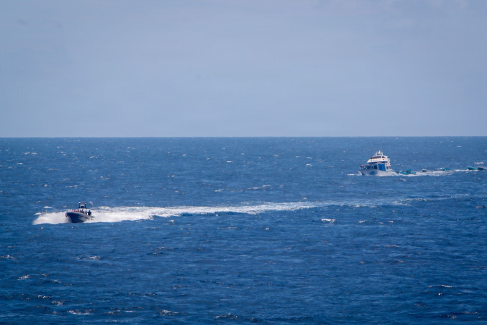 The image shows two vessels on open ocean waters under a hazy sky. A smaller, fast-moving rigid inflatable boat (RIB) on the left is generating a significant white wake, appearing to pursue or intercept a larger vessel on the right. The sea conditions appear moderately choppy with visible wave action.
