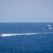 The image shows two vessels on open ocean waters under a hazy sky. A smaller, fast-moving rigid inflatable boat (RIB) on the left is generating a significant white wake, appearing to pursue or intercept a larger vessel on the right. The sea conditions appear moderately choppy with visible wave action.