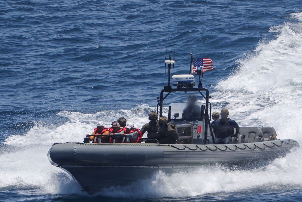 A rigid-hull inflatable boat (RHIB) is underway at high speed on open ocean waters. The vessel carries several personnel, some wearing orange life vests and others in military/tactical gear, suggesting a rescue or interdiction operation. An American flag is mounted on the boat's center console mast.