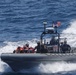 A rigid-hull inflatable boat (RHIB) is underway at high speed on open ocean waters. The vessel carries several personnel, some wearing orange life vests and others in military/tactical gear, suggesting a rescue or interdiction operation. An American flag is mounted on the boat's center console mast.