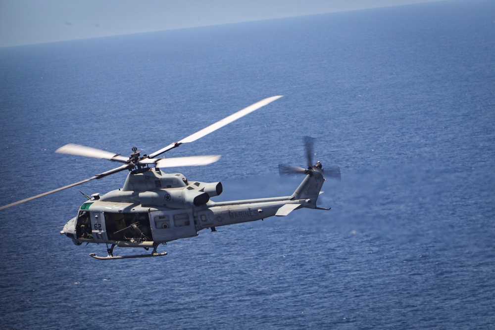 A U.S. Marines military helicopter, identified by the 'MARINES' text on its fuselage and tail number '62', is captured in flight over open ocean. The aircraft appears to be a UH-1Y Venom (Huey) helicopter, painted in standard military gray. The image is taken from an aerial perspective, likely from another aircraft flying in formation.
