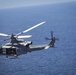 A U.S. Marines military helicopter, identified by the 'MARINES' text on its fuselage and tail number '62', is captured in flight over open ocean. The aircraft appears to be a UH-1Y Venom (Huey) helicopter, painted in standard military gray. The image is taken from an aerial perspective, likely from another aircraft flying in formation.