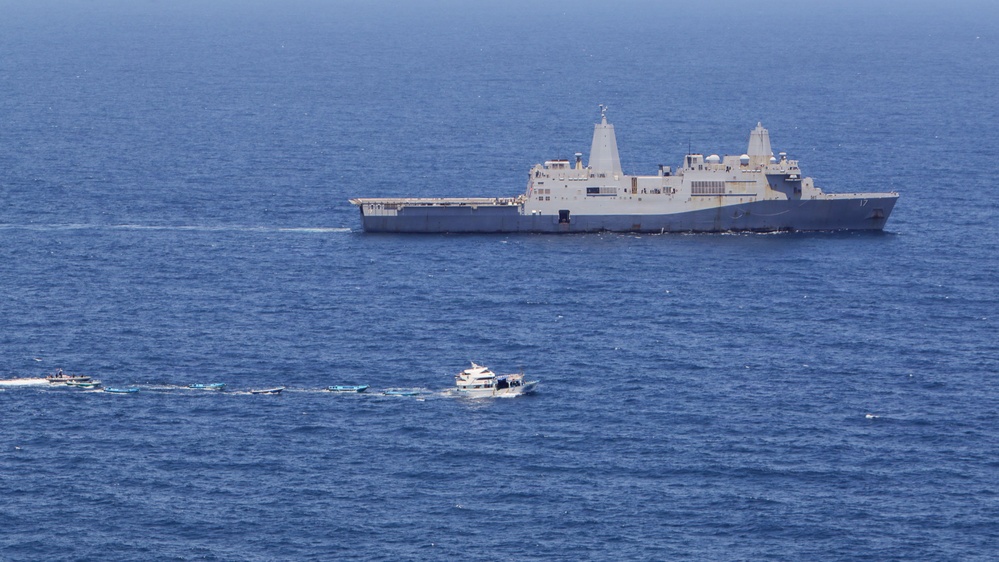 A large gray naval warship, bearing the hull number '17', is anchored or underway in open blue ocean waters. Several smaller civilian and military vessels are visible in the foreground, appearing to approach or transit near the large ship. The scene suggests a maritime interaction or escort situation in calm sea conditions.