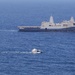 A large gray naval warship, bearing the hull number '17', is anchored or underway in open blue ocean waters. Several smaller civilian and military vessels are visible in the foreground, appearing to approach or transit near the large ship. The scene suggests a maritime interaction or escort situation in calm sea conditions.