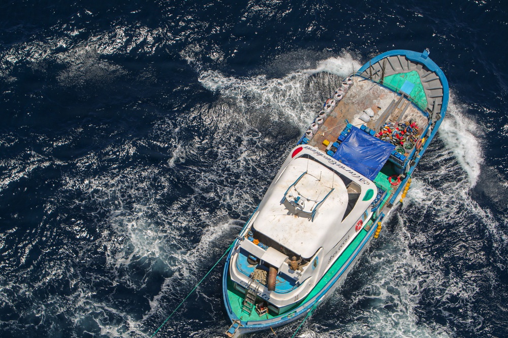 An aerial photograph taken from above shows a small to medium-sized vessel moving through deep blue open water, leaving a white wake trail. The boat appears to be a fishing or utility vessel with a white cabin structure at the bow and a crowded rear deck covered with a blue tarp and various colorful items. The vessel has a distinctive blue and green hull and appears to be traveling at moderate speed.