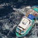 An aerial photograph taken from above shows a small to medium-sized vessel moving through deep blue open water, leaving a white wake trail. The boat appears to be a fishing or utility vessel with a white cabin structure at the bow and a crowded rear deck covered with a blue tarp and various colorful items. The vessel has a distinctive blue and green hull and appears to be traveling at moderate speed.