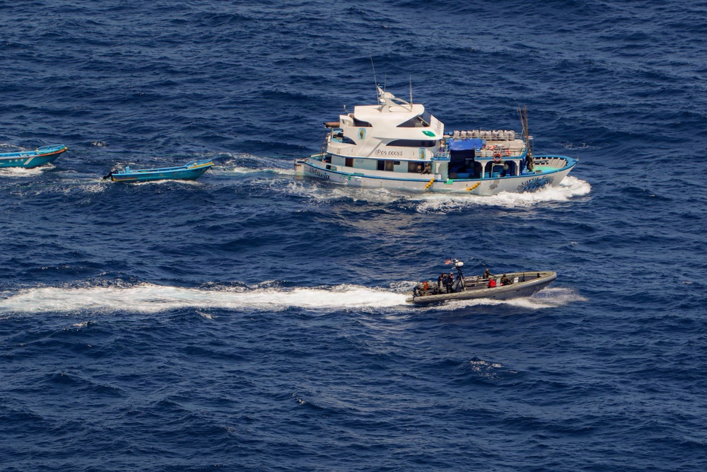 An aerial photograph shows a maritime interdiction operation in open ocean waters. A larger white and blue multi-deck vessel is being approached by a rigid inflatable boat (RIB) carrying what appear to be law enforcement or military personnel. Two smaller blue boats are also visible in the scene, one appearing to be towed behind the larger vessel.