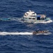 An aerial photograph shows a maritime interdiction operation in open ocean waters. A larger white and blue multi-deck vessel is being approached by a rigid inflatable boat (RIB) carrying what appear to be law enforcement or military personnel. Two smaller blue boats are also visible in the scene, one appearing to be towed behind the larger vessel.