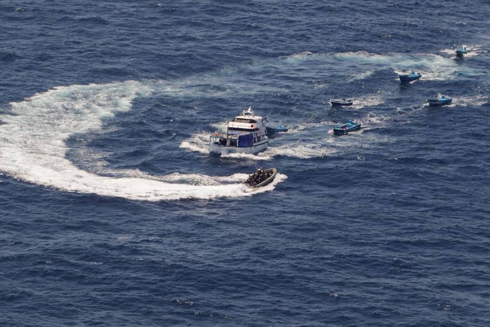 An aerial photograph shows a maritime interdiction or law enforcement operation in open ocean. A larger white motor vessel is surrounded by multiple smaller boats, with one rigid inflatable boat (RIB) carrying what appears to be a tactical team approaching the larger vessel. The scene shows active maneuvering with visible wake patterns from multiple craft.