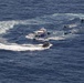 An aerial photograph shows a maritime interdiction or law enforcement operation in open ocean. A larger white motor vessel is surrounded by multiple smaller boats, with one rigid inflatable boat (RIB) carrying what appears to be a tactical team approaching the larger vessel. The scene shows active maneuvering with visible wake patterns from multiple craft.