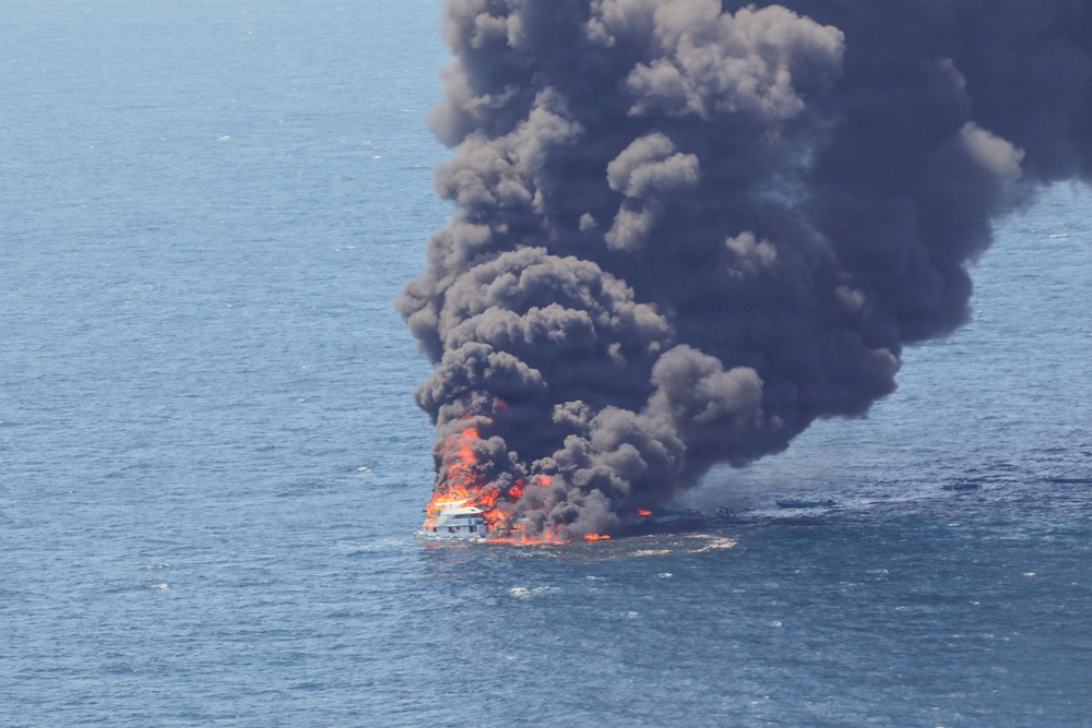 An aerial photograph shows a vessel engulfed in intense flames on open water. Large billowing columns of dark black smoke rise high into the sky, indicating a significant fire involving fuel or petroleum-based materials. The vessel appears to be a mid-sized motorboat or yacht, largely consumed by the fire.