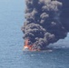 An aerial photograph shows a vessel engulfed in intense flames on open water. Large billowing columns of dark black smoke rise high into the sky, indicating a significant fire involving fuel or petroleum-based materials. The vessel appears to be a mid-sized motorboat or yacht, largely consumed by the fire.