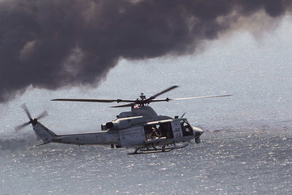 A U.S. Marines military helicopter, identifiable as a UH-1Y Venom (tail number 62), is flying low over a body of water. The helicopter has its side door open with what appears to be a crew member or door gunner visible. Dark smoke clouds are visible in the background, suggesting a nearby fire or exercise activity.