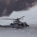 A U.S. Marines military helicopter, identifiable as a UH-1Y Venom (tail number 62), is flying low over a body of water. The helicopter has its side door open with what appears to be a crew member or door gunner visible. Dark smoke clouds are visible in the background, suggesting a nearby fire or exercise activity.
